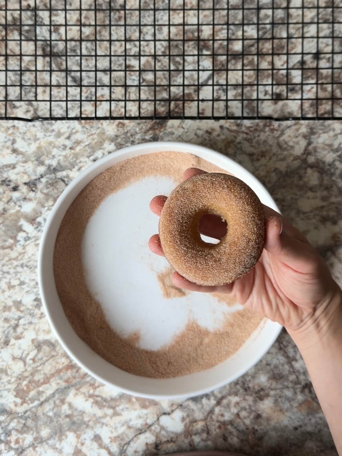 A baked sourdough apple cider donut being dipped in a mixture of cinnamon sugar.