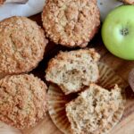 Sourdough apple muffins with cinnamon streusel topping on a wooden cutting board with one green apple amongst them.