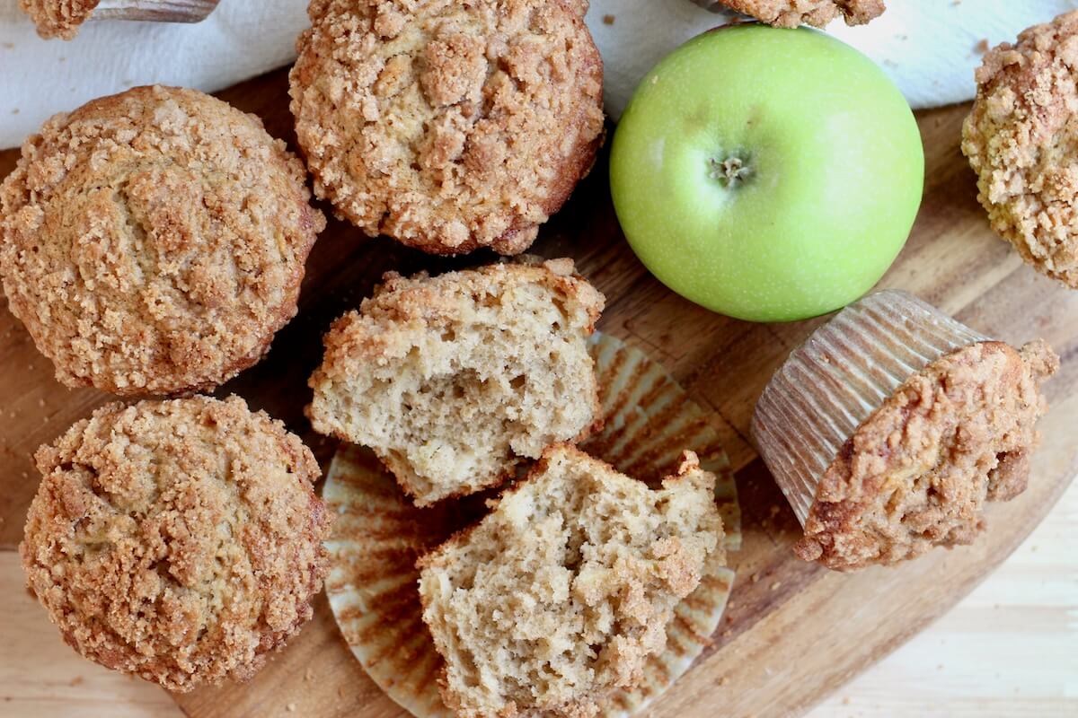 Sourdough apple muffins with cinnamon streusel topping on a wooden cutting board with one green apple amongst them.
