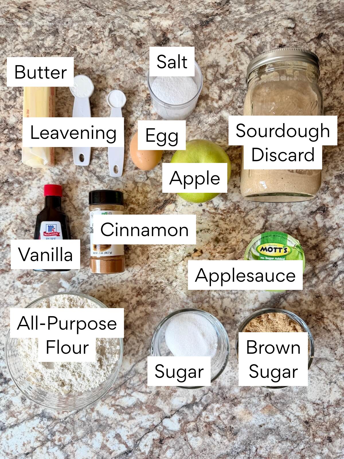 The ingredients to make sourdough apple muffins. Each ingredient is labeled with text. They include butter, leavening, salt, egg, apple, sourdough discard, vanilla, cinnamon, applesauce, all-purpose flour, sugar, and brown sugar.