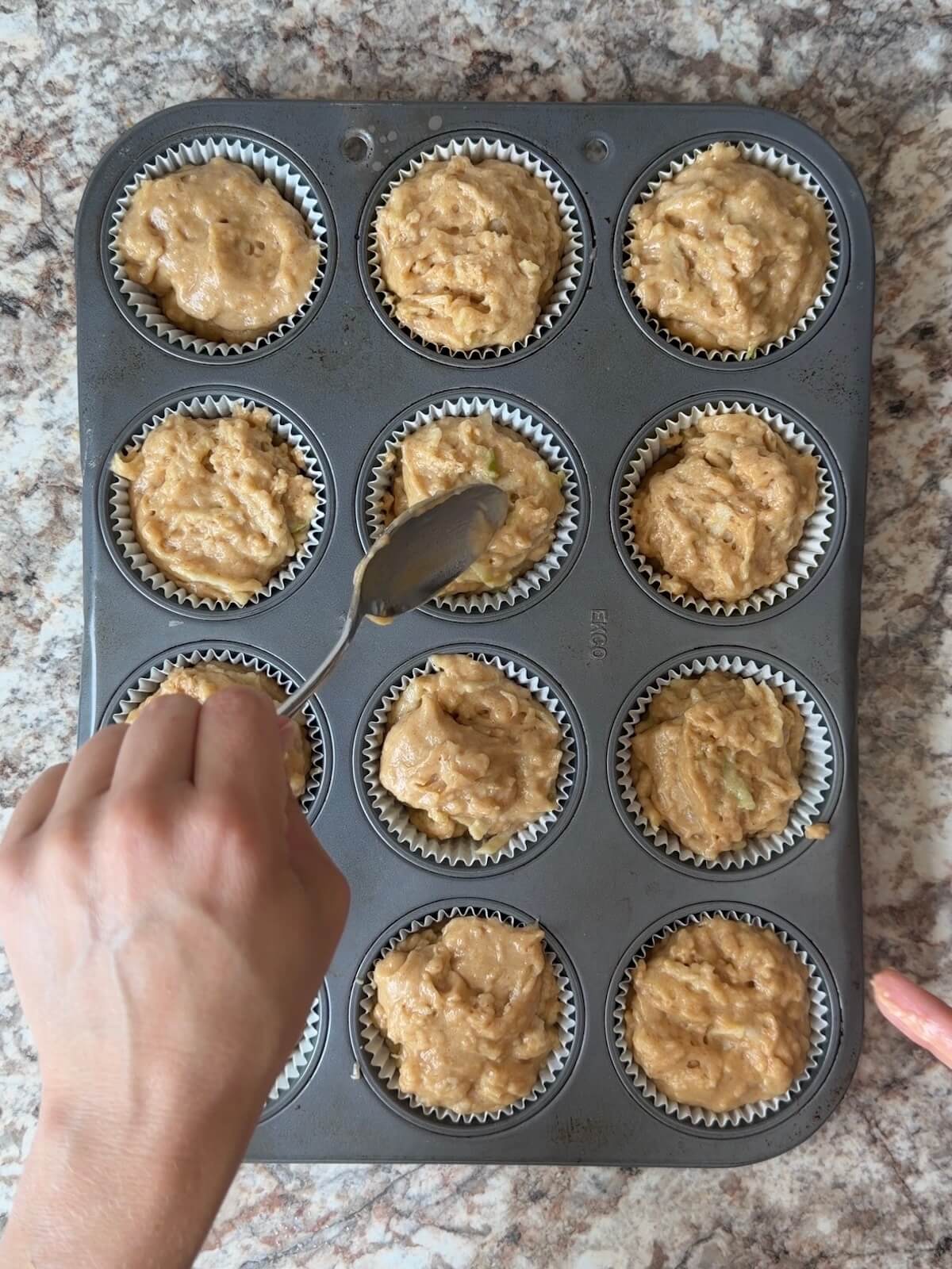 A paper-lined muffin tin being filled with sourdough apple muffin batter.