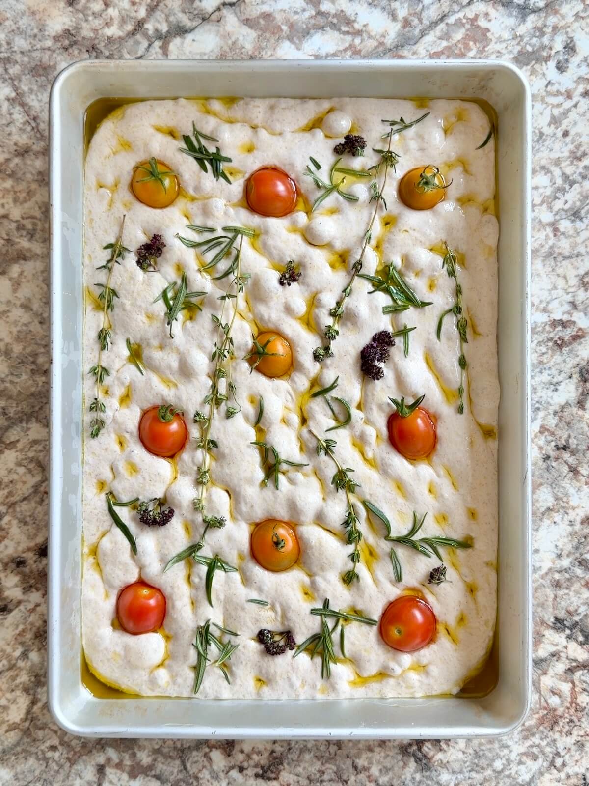 Proofed sourdough discard focaccia dough topped with herbs and cherry tomatoes.