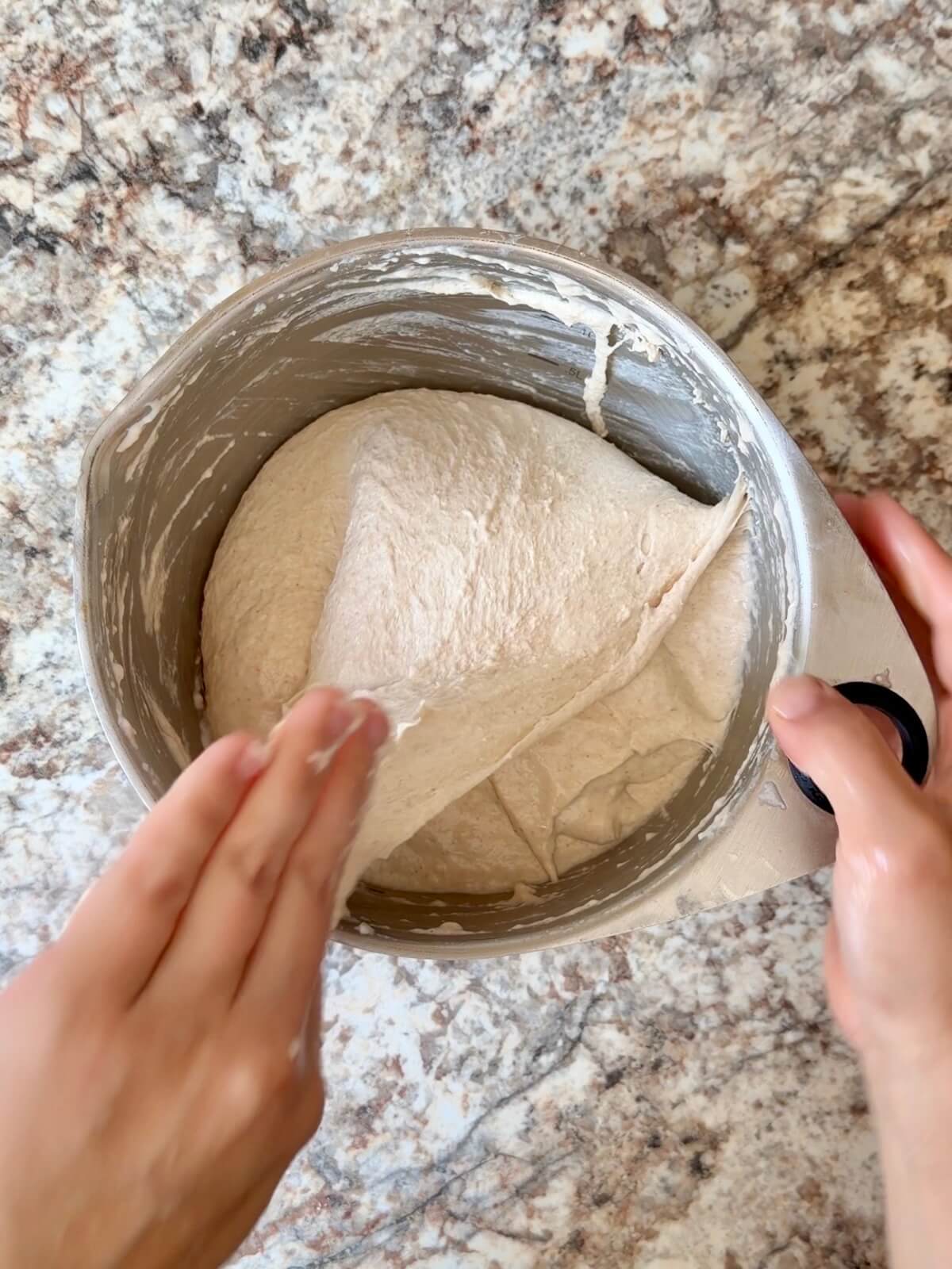 Two hands stretching and folding sourdough focaccia bread dough in a bowl.