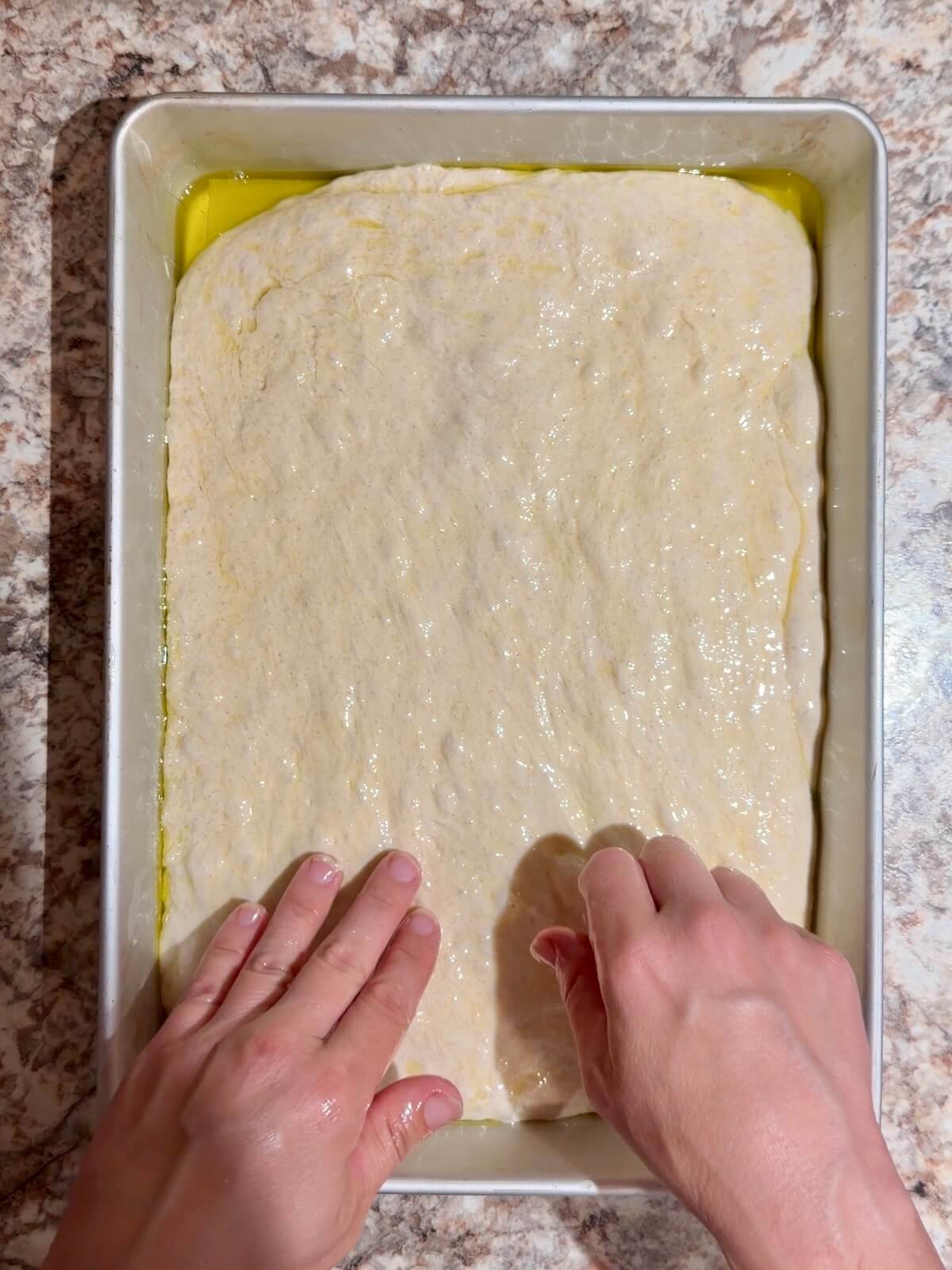 Two hands spreading fermented sourdough focaccia dough into an oil-coated baking pan.