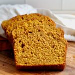 Slices of sourdough discard pumpkin bread on a wooden cutting board with white and orange cloth napkins in the background.