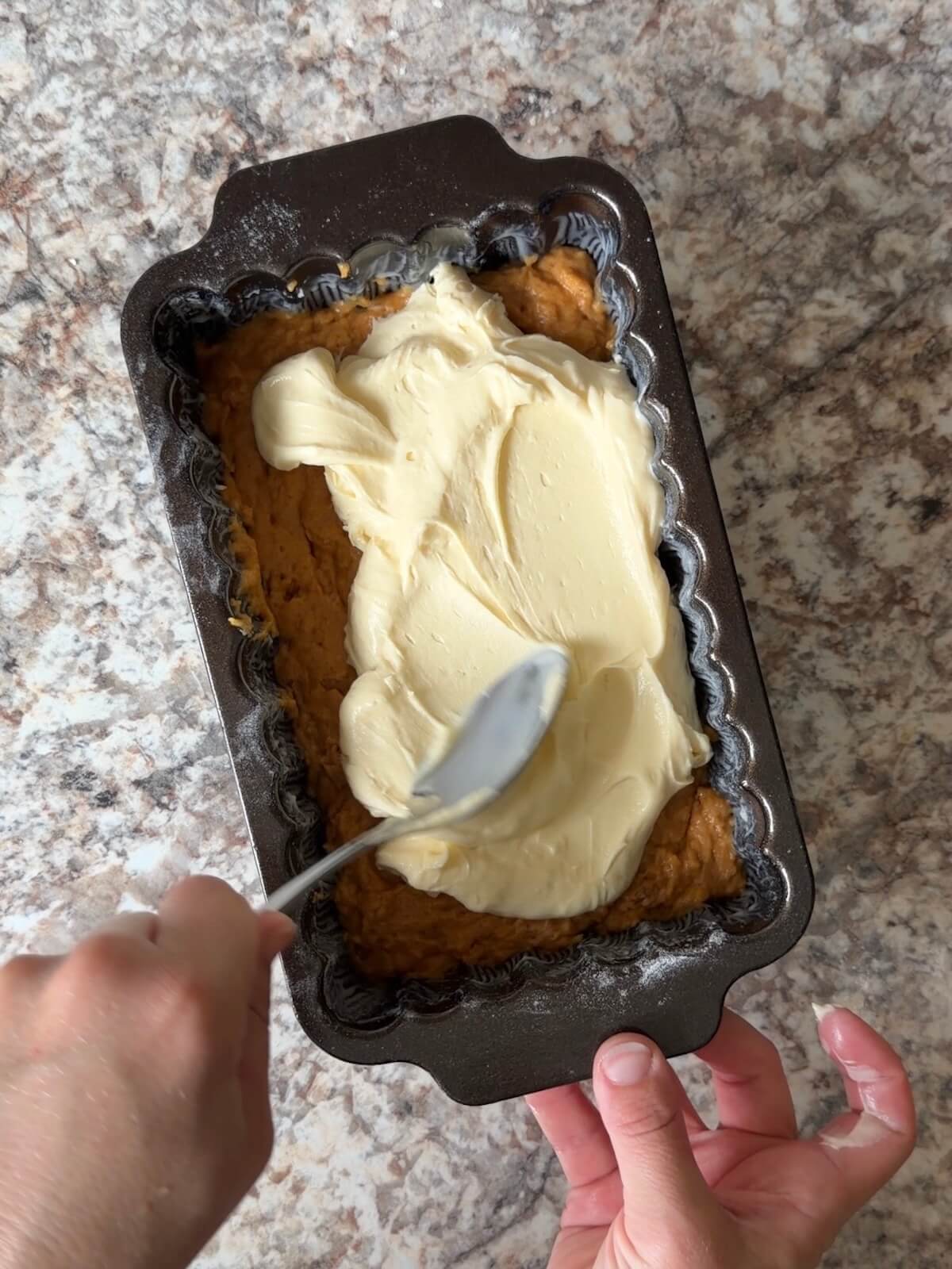 The cream cheese layer being spread on top of some of the pumpkin bread batter in a loaf pan.