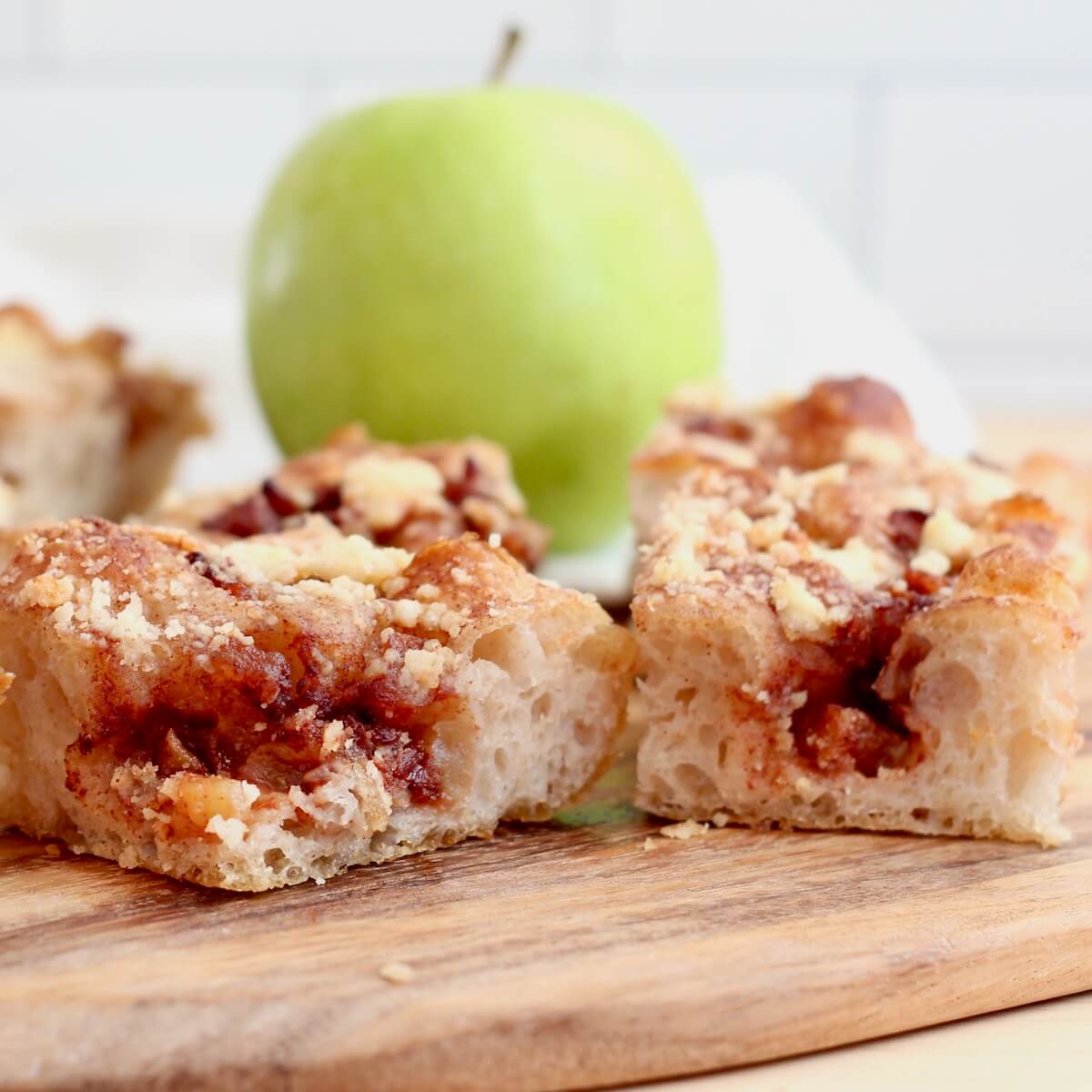 Several pieces of apple pie sourdough focaccia on a cutting board. There is a green apple out of focus in the background.