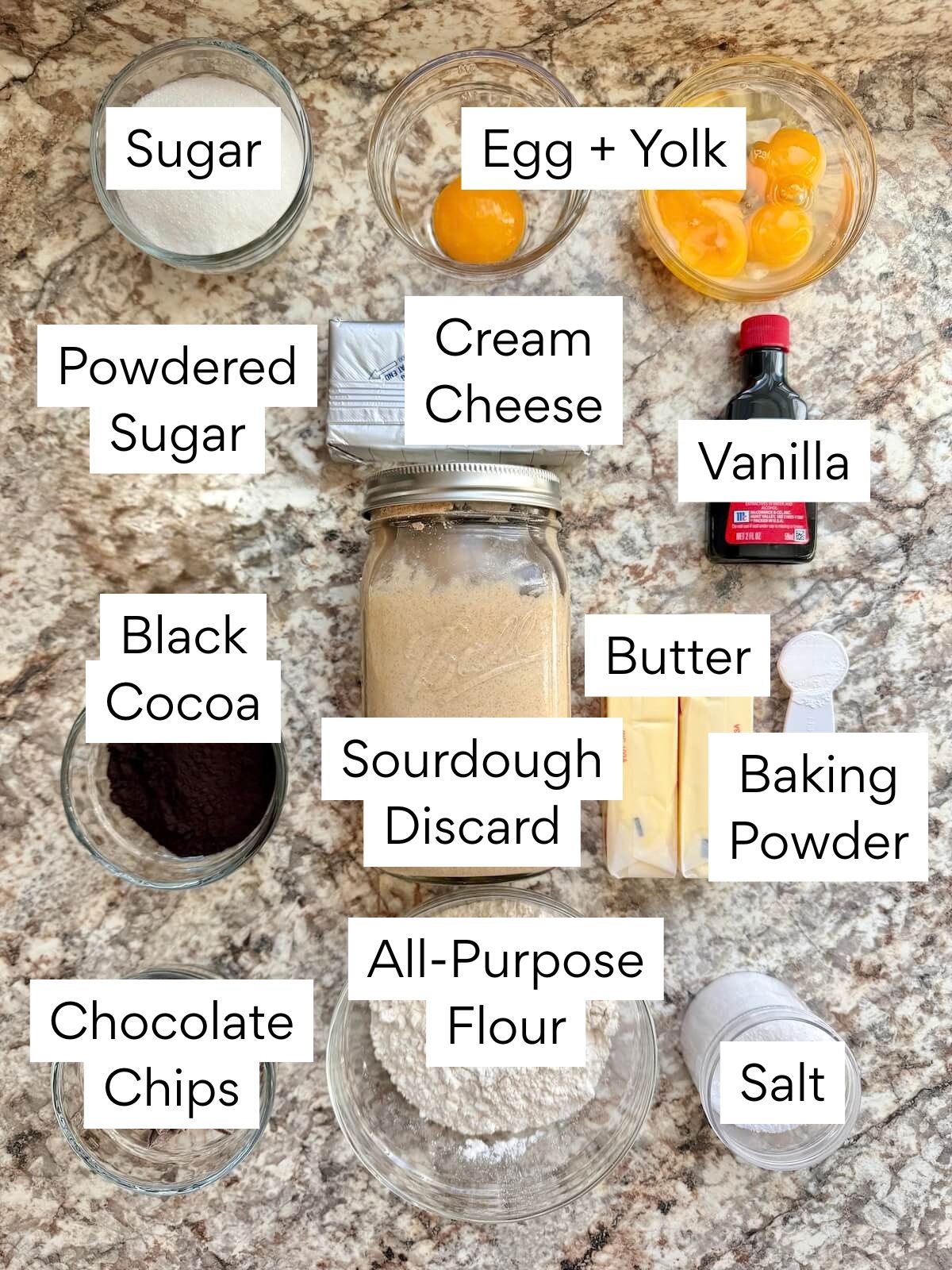 The ingredients to make black cocoa sourdough brownies. Each ingredient is labeled with text. They include sugar, egg, egg yolk, powdered sugar, cream cheese, vanilla, black cocoa, sourdough discard, butter, baking powder, chocolate chips, all-purpose flour, and salt.