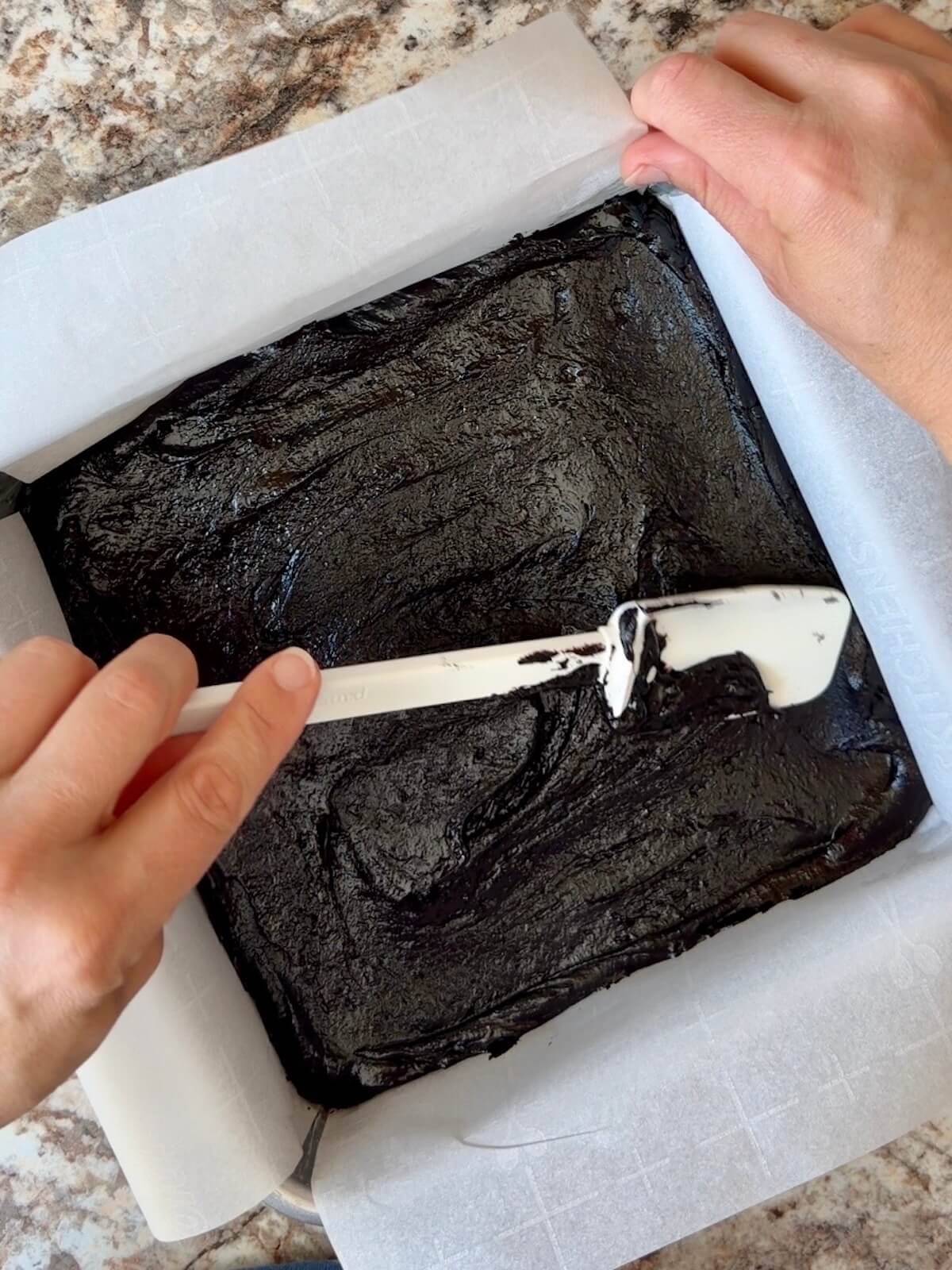 Black cocoa sourdough brownie batter being spread in a parchment-lined square baking pan.