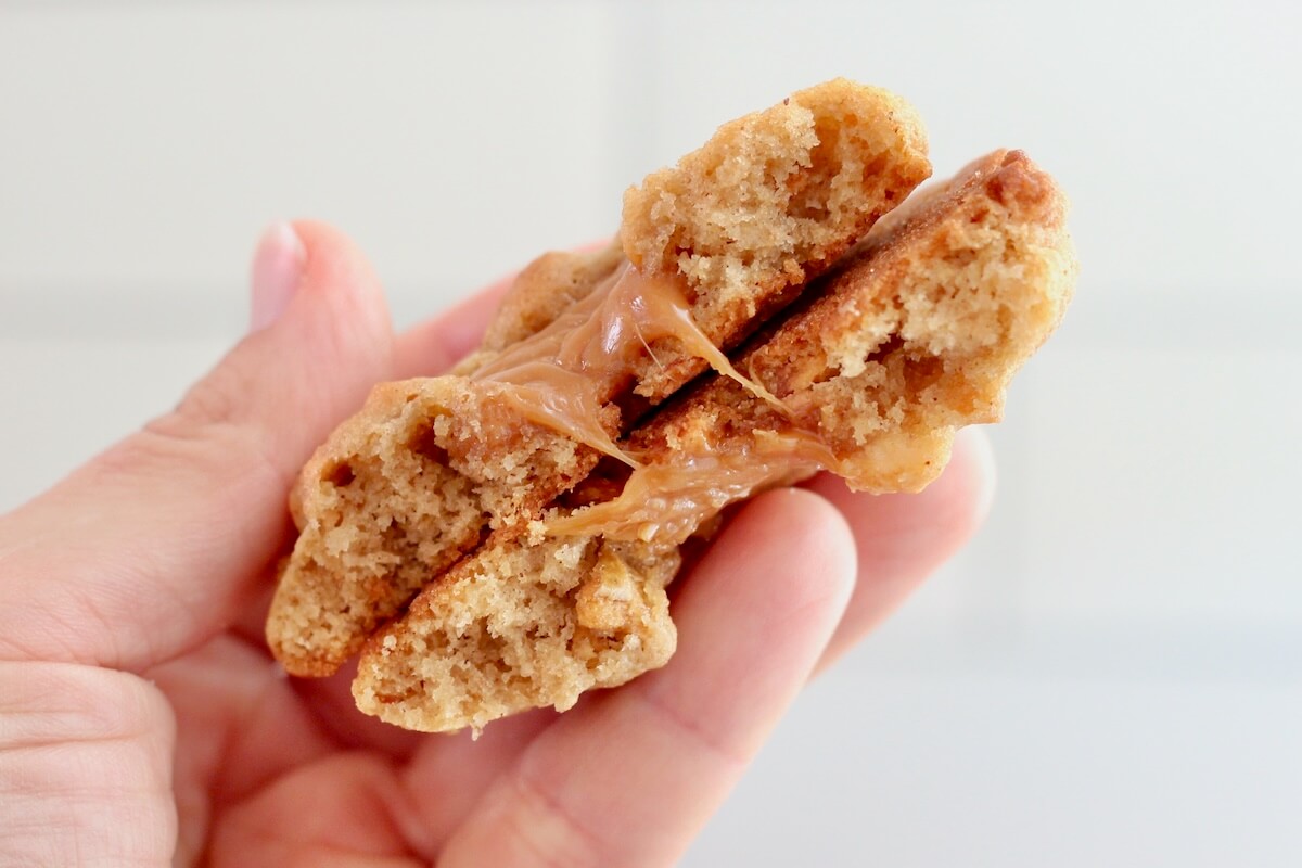 A hand holding a caramel apple sourdough cookie that's been split in half to show the melty caramel-filled center.