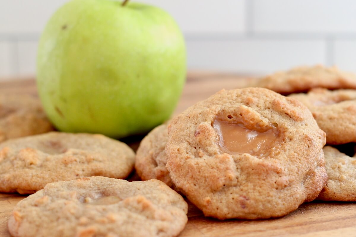 Several caramel apple sourdough cookies on a cutting board with a fresh green apple out of focus in the background.
