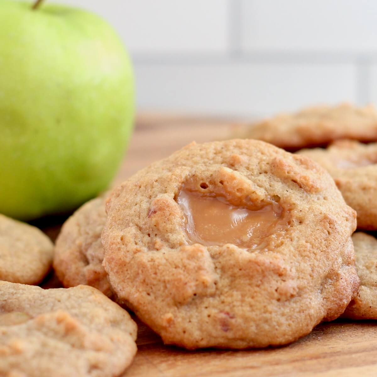 Several caramel apple sourdough cookies on a cutting board with a fresh green apple out of focus in the background.