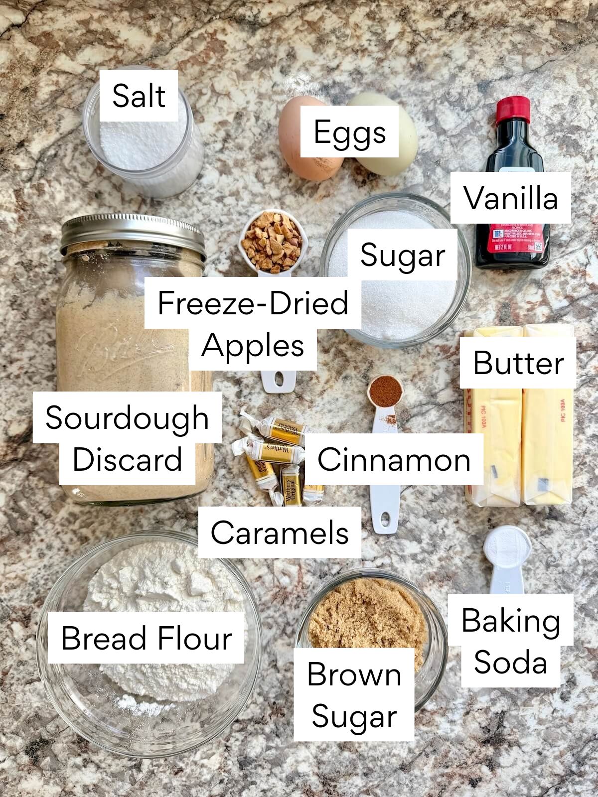 The ingredients to make sourdough caramel apple cookies. Each ingredient is labeled with text. They include salt, eggs, vanilla, sugar, freeze-dried apples, sourdough discard, caramels, cinnamon, butter, bread flour, brown sugar, and baking soda.