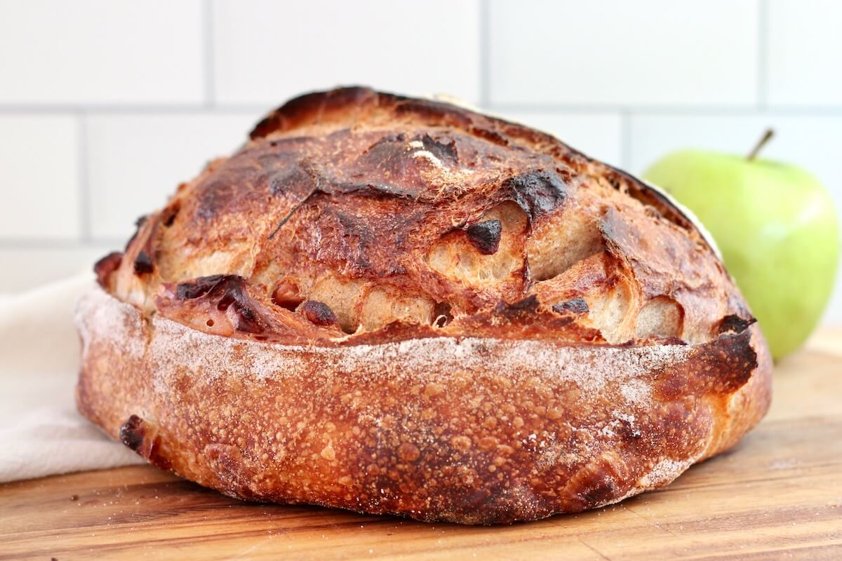 A whole loaf of apple cinnamon sourdough bread on a wooden cutting board with a fresh green apple out of focus in the background.