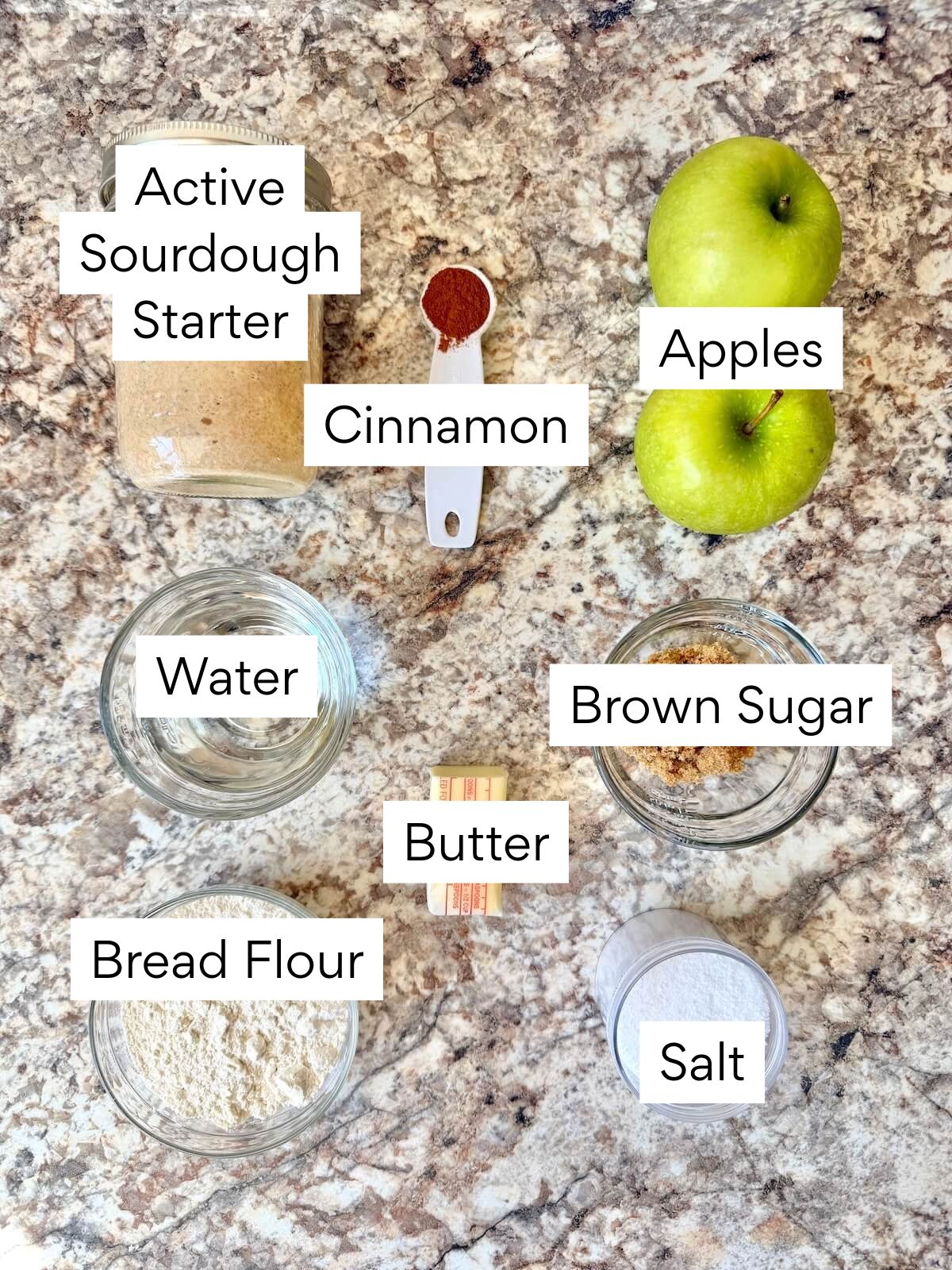 The ingredients to make cinnamon apple sourdough bread. Each ingredient is labeled with text. They include active sourdough starter, cinnamon, apples, water, butter, brown sugar, bread flour, and salt.