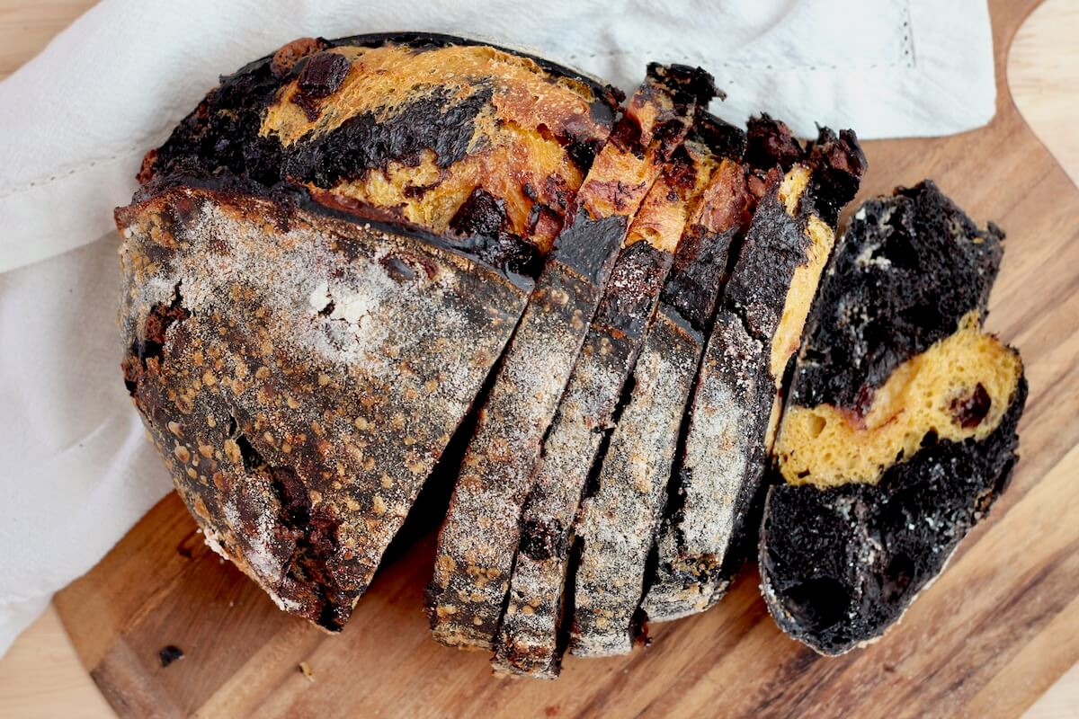 A loaf of black and orange sourdough bread sliced on a wooden cutting board.