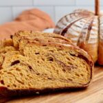 Slices of pumpkin spice sourdough bread on a wooden cutting board. There is a whole loaf of pumpkin-shaped bread out of focus in the background.