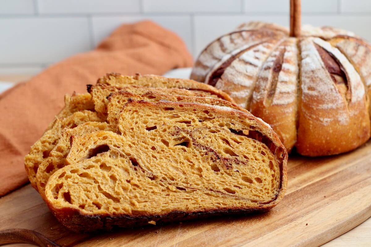Slices of pumpkin spice sourdough bread on a wooden cutting board. There is a whole loaf of pumpkin-shaped bread out of focus in the background.