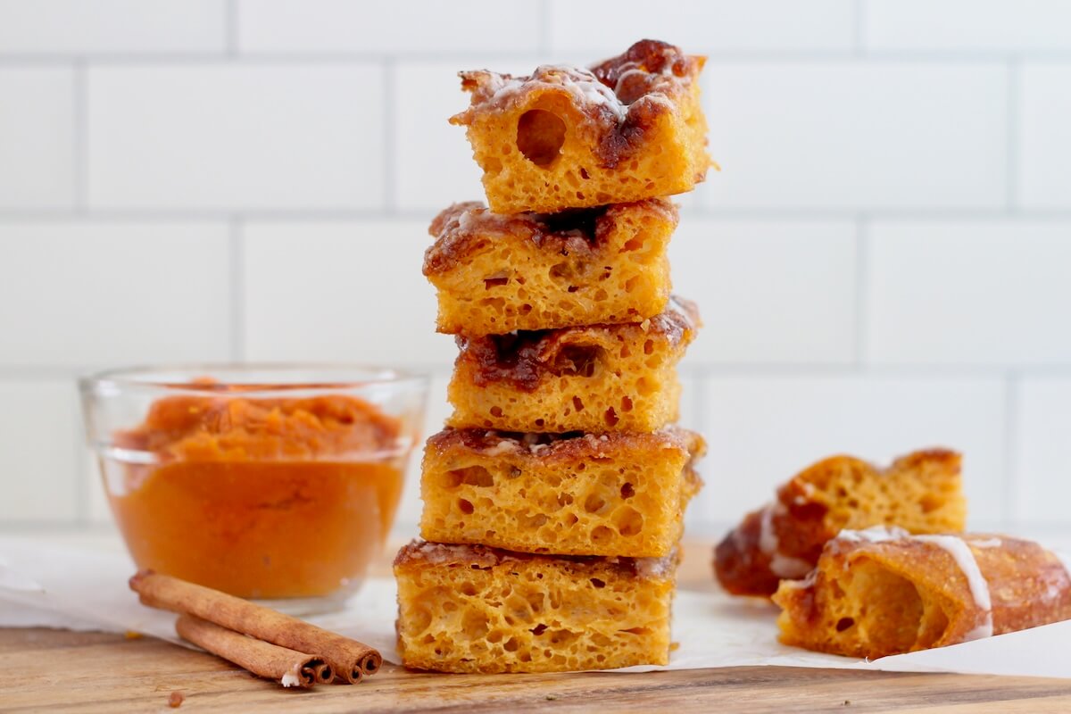 A stack of five pieces of pumpkin spice sourdough focaccia on a cutting board with a small bowl of pumpkin, two cinnamon sticks, and two more pieces of focaccia out of focus in the background.