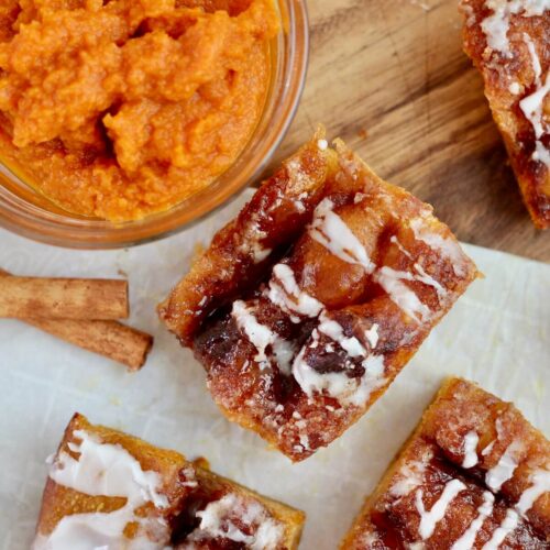 Pieces of sweet pumpkin sourdough focaccia on a wooden cutting board. There is a small bowl of pumpkin puree and two cinnamon sticks on the board with the bread.