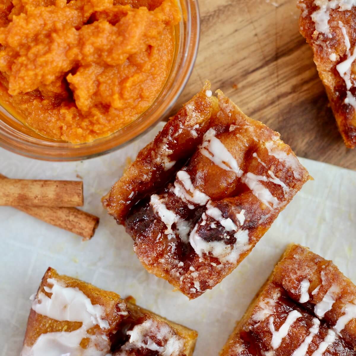 Pieces of sweet pumpkin sourdough focaccia on a wooden cutting board. There is a small bowl of pumpkin puree and two cinnamon sticks on the board with the bread.