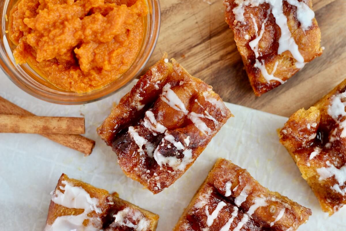 Pieces of sweet pumpkin sourdough focaccia on a wooden cutting board. There is a small bowl of pumpkin puree and two cinnamon sticks on the board with the bread.