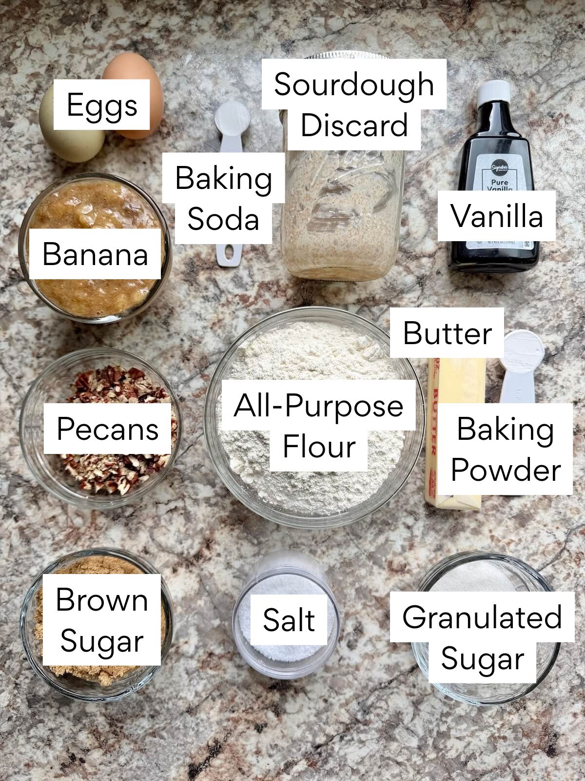 The ingredients to make sourdough banana bread. Each ingredient is labeled with text. They include eggs, banana, pecans, brown sugar, sourdough discard, baking soda, all-purpose flour, salt, granulated sugar, baking powder, butter, and vanilla.