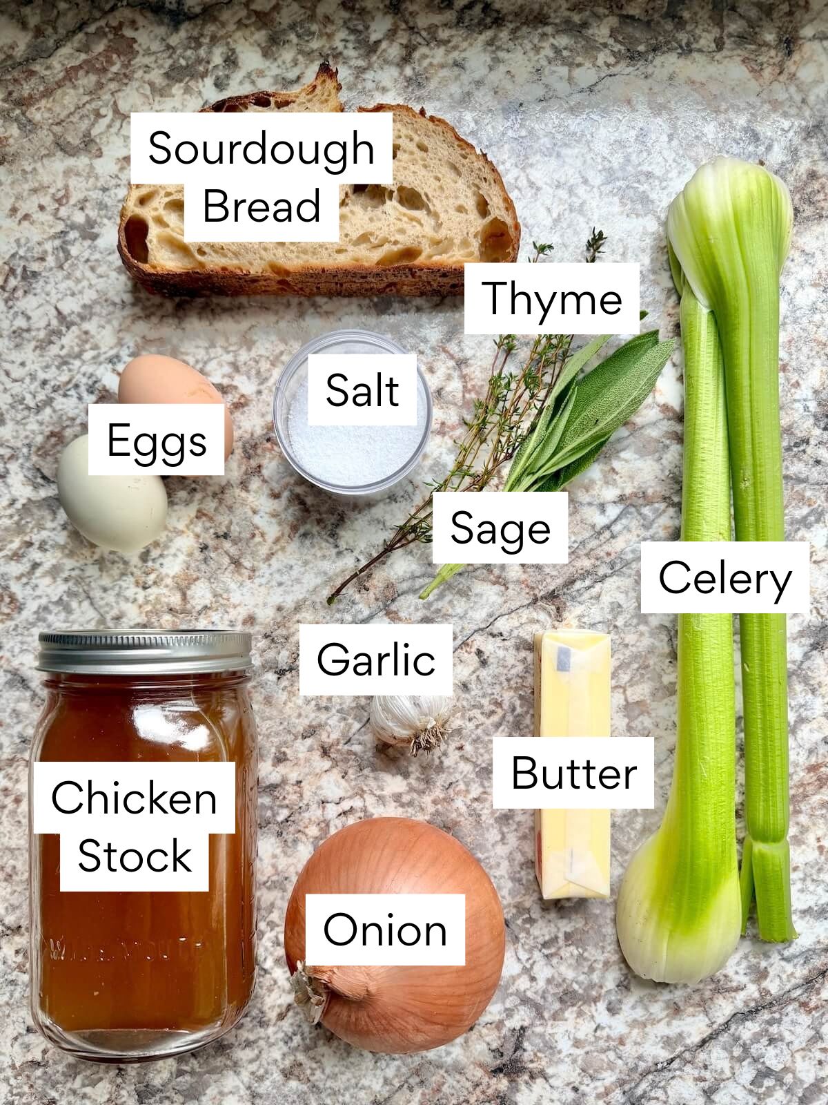 The ingredients to make sourdough bread stuffing. Each ingredient is labeled with text. They include sourdough bread, eggs, salt, thyme, sage, celery, garlic, chicken stock, butter, and onion.