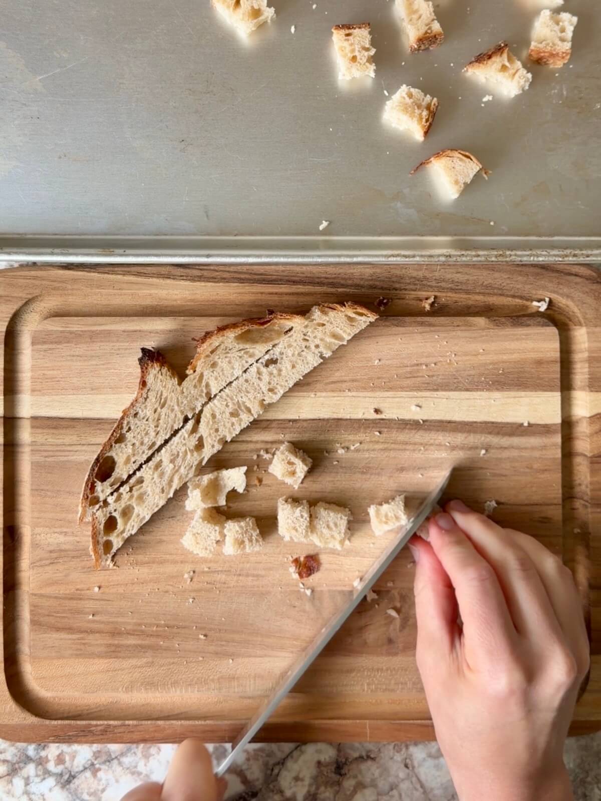 Sourdough bread being cut into small cubes on a wooden cutting board.