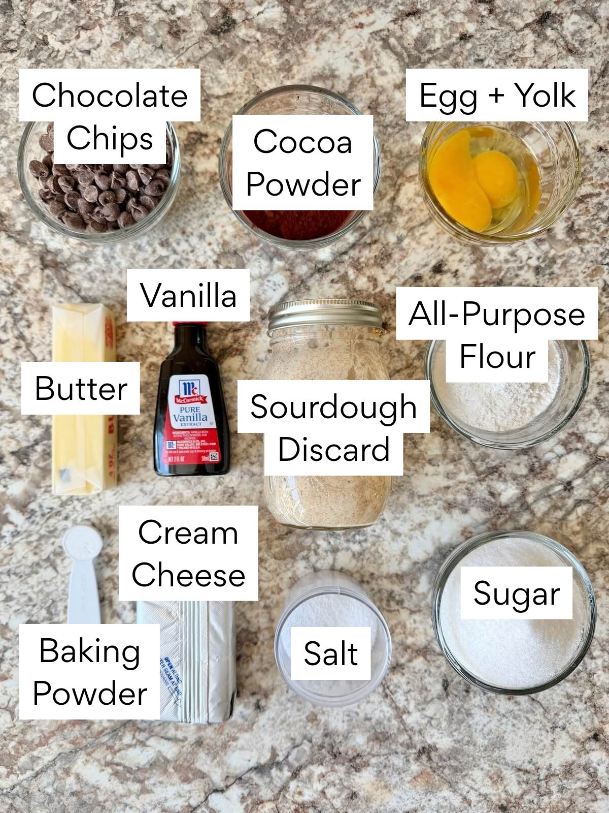 The ingredients to make sourdough cheesecake brownies. Each ingredient is labeled with text. They include chocolate chips, cocoa powder, egg and egg yolk, butter, vanilla, sourdough discard, all-purpose flour, baking powder, cream cheese, salt, and sugar.