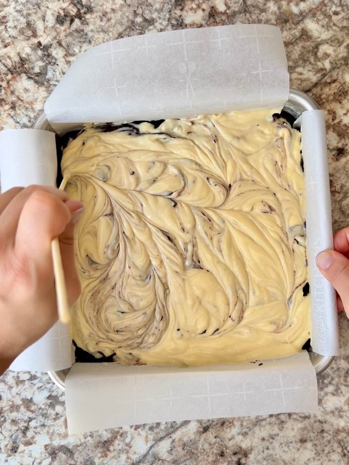 The cheesecake layer being swirled into the brownie batter in a square pan.