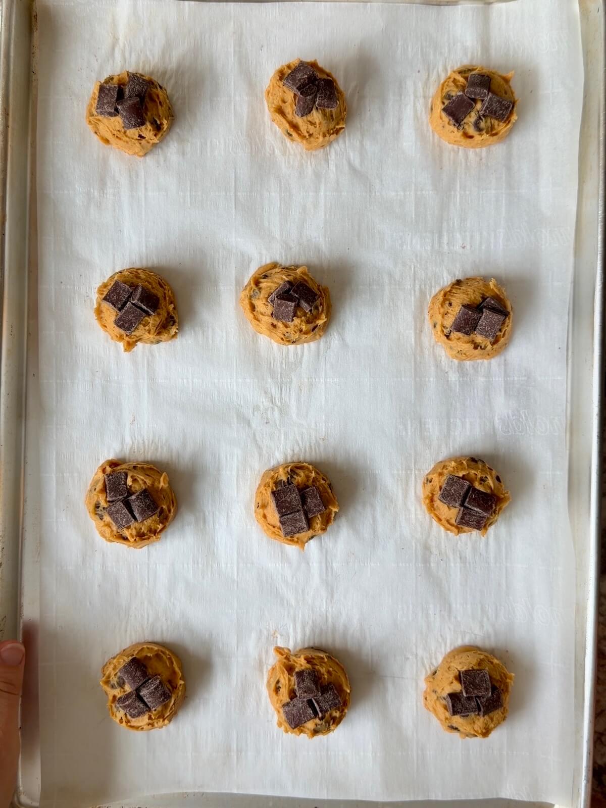 Twelve cookie dough balls with extra chocolate pressed on top on a parchment-lined baking sheet.