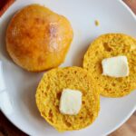 Two sourdough pumpkin dinner rolls on a small white plate. One roll is split in half and each half has a pat of butter on it.