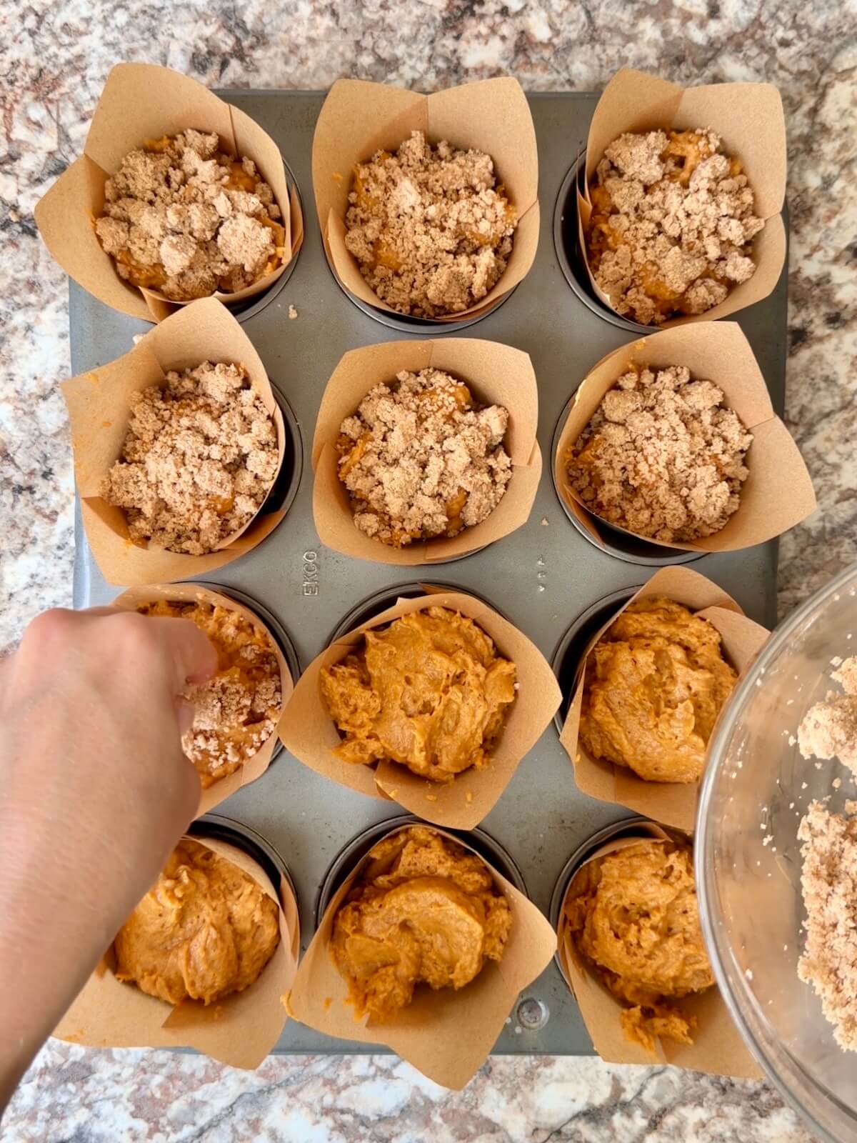 Parchment lined muffin pan filled with sourdough pumpkin muffin batter. Each muffin is being topped with cinnamon streusel.