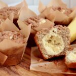 Sourdough discard banana muffins with cinnamon streusel on a wooden cutting board. One of the muffins is split in half to show the cream cheese filling.