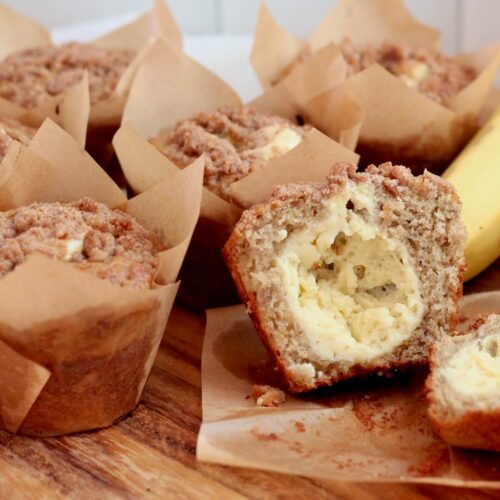 Sourdough discard banana muffins with cinnamon streusel on a wooden cutting board. One of the muffins is split in half to show the cream cheese filling.