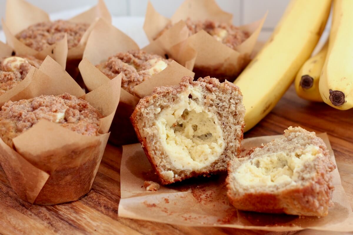 Sourdough discard banana muffins with cinnamon streusel on a wooden cutting board. One of the muffins is split in half to show the cream cheese filling.