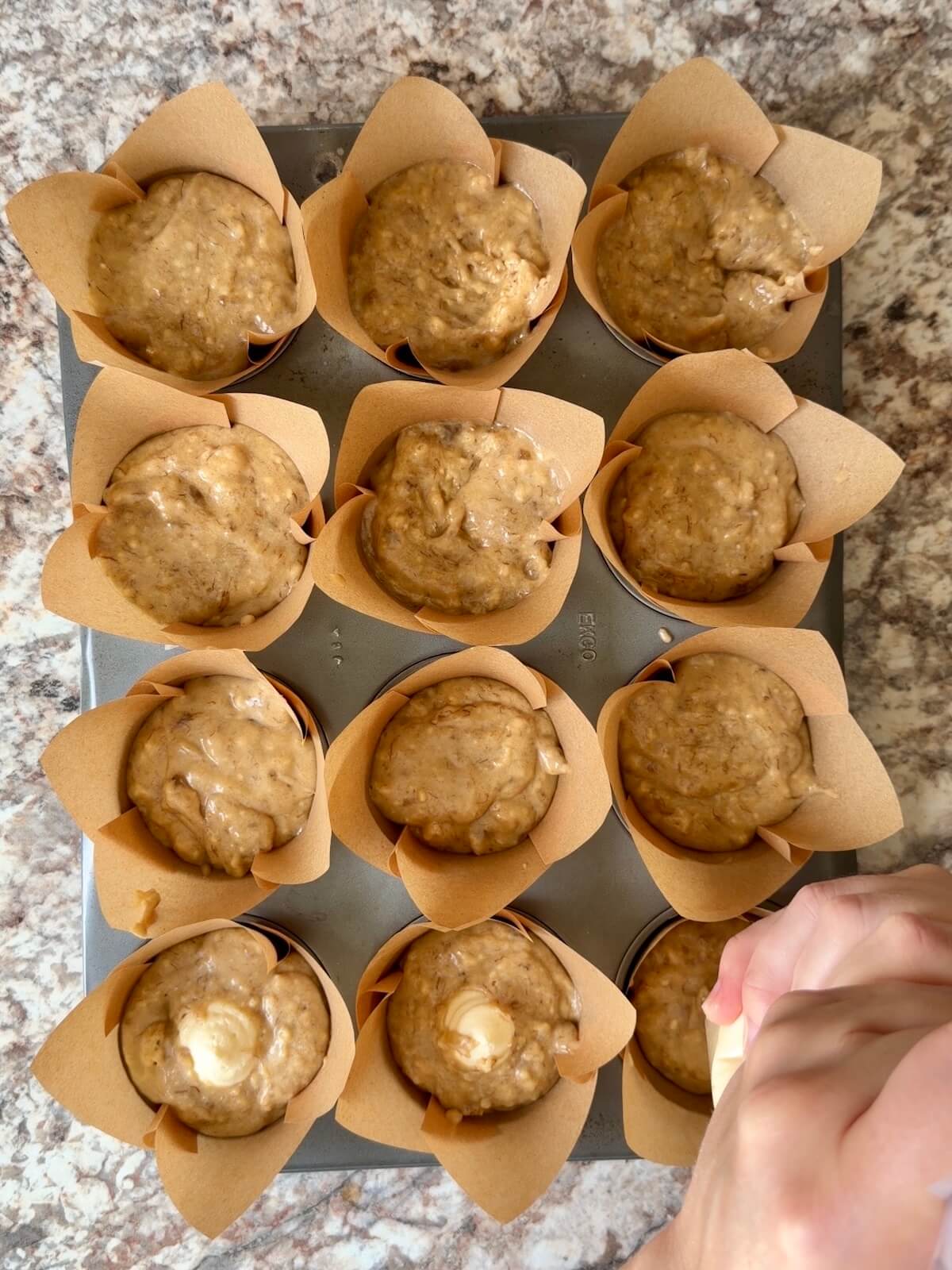 Sourdough banana muffin batter in a paper lined muffin tin. The muffins are being piped with cream cheese filling.
