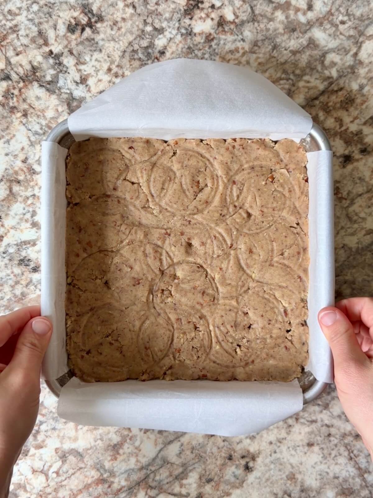 Pecan cookie dough pressed into the bottom of a square, parchment-lined baking pan.