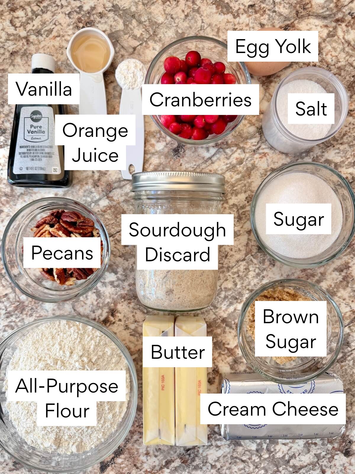 The ingredients to make sourdough cranberry pecan cheesecake bars. Each ingredient is labeled with text. They include vanilla, orange juice, cranberries, egg yolk, salt, pecans, sourdough discard, sugar, all-purpose flour, butter, brown sugar, and cream cheese.