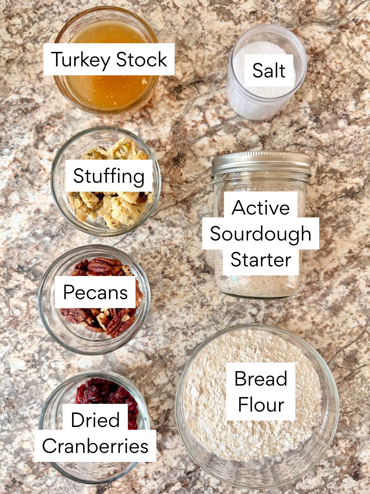 The ingredients to make Thanksgiving stuffing sourdough bread. Each ingredient is labeled with text. They include turkey stock, stuffing, pecans, dried cranberries, salt, active sourdough starter, and bread flour.